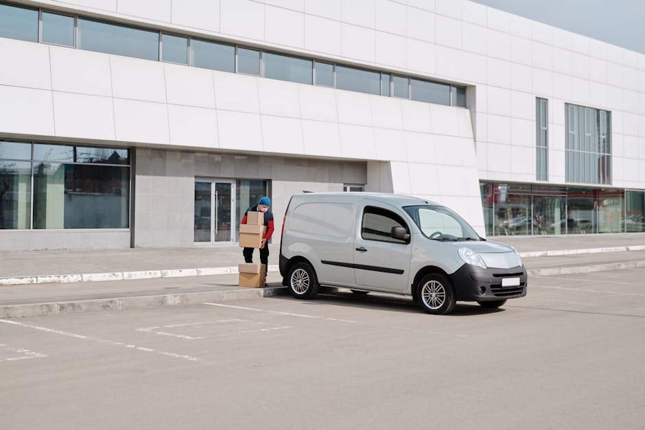 A person wearing a red jacket and black trousers is standing outside a modern, white multi-story building with large glass windows, loading three cardboard boxes into the back of a small, grey panel van parked on a paved lot. The boxes are stacked one on top of another, and the person appears to be placing or retrieving them from the vehicle. The van is positioned near the building entrance, which has a glass door, and the scene is well-lit with natural daylight. The parking area is marked with white lines, and there are no other vehicles or people visible nearby. This image depicts the typical process of packing and loading belongings during a home relocation or moving service, which might be provided by Man with Van Addiscombe, aligned with the parking and loading rules for movers in Addiscombe.