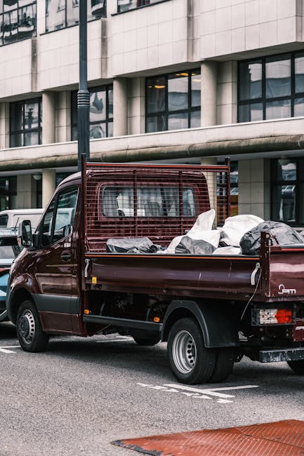 A brown flatbed van parked on a city street in front of a modern office building with large glass windows, loaded with various used furniture and household items wrapped in plastic and fabric, including a white cushion and a cardboard box, ready for transportation or disposal. The van has a metal cargo barrier behind the cab for safety, and there is a lamppost standing nearby. The scene depicts a typical home relocation or collection process, with the vehicle prepared for furniture transport or bulky waste pickup, reflecting services offered by Man with Van Addiscombe in the context of house removals and moving logistics.