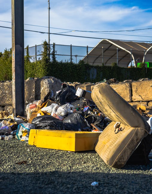 A pile of mixed household waste outside a property, including black and white plastic rubbish bags, a yellow plastic storage container, an old fabric-upholstered armchair turned on its side, and various discarded items scattered on a gravel surface. Behind the waste, there is a stone wall, a wooden fence, and utility poles with overhead wires, indicating an outdoor setting in a residential or semi-industrial area. The scene is illuminated by daylight, suggesting clear weather. This image depicts the type of bulky waste removal often handled during home relocation or furniture transport services, as provided by Man with Van Addiscombe, highlighting the importance of proper waste disposal during moving and clearance processes.