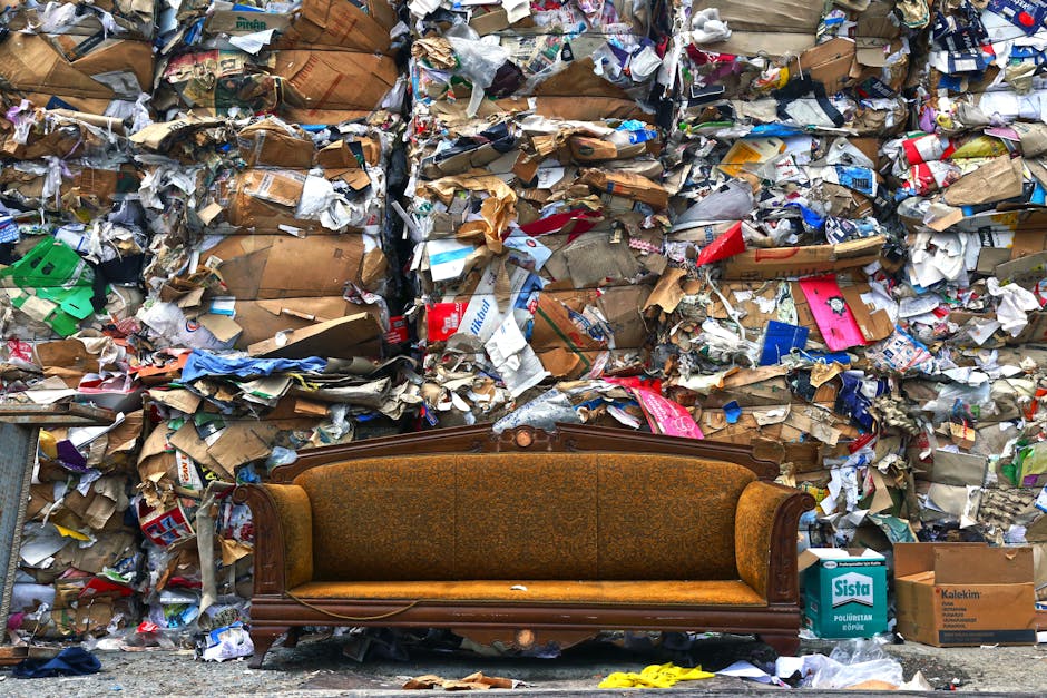A brown flatbed van parked on a city street in front of a modern office building with large glass windows, loaded with various used furniture and household items wrapped in plastic and fabric, including a white cushion and a cardboard box, ready for transportation or disposal. The van has a metal cargo barrier behind the cab for safety, and there is a lamppost standing nearby. The scene depicts a typical home relocation or collection process, with the vehicle prepared for furniture transport or bulky waste pickup, reflecting services offered by Man with Van Addiscombe in the context of house removals and moving logistics.