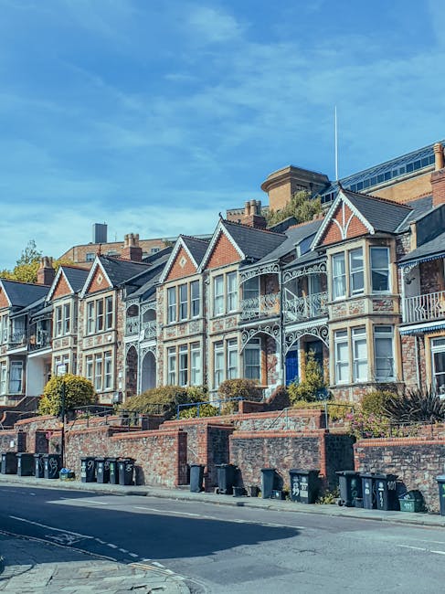 A row of Victorian-style terraced houses with ornate white metal balconies and bay windows situated behind a brick wall with small landscaped gardens, located on a residential street during daylight hours under a partly cloudy sky. In the foreground, there are multiple black wheelie bins lined along the pavement, indicating local waste collection, with a dark grey van parked nearby, possibly used for home relocation or furniture transport by Man with Van Addiscombe. The scene captures the typical urban setting for house removals and moving services, illustrating a professional environment for loading and unloading possessions during a house move or residential clearance, with the houses' architecture suggesting an older, established neighbourhood in the Croydon area. The presence of clear weather, well-maintained gardens, and accessible street features highlights the typical logistical environment for professional removals in London suburbs.