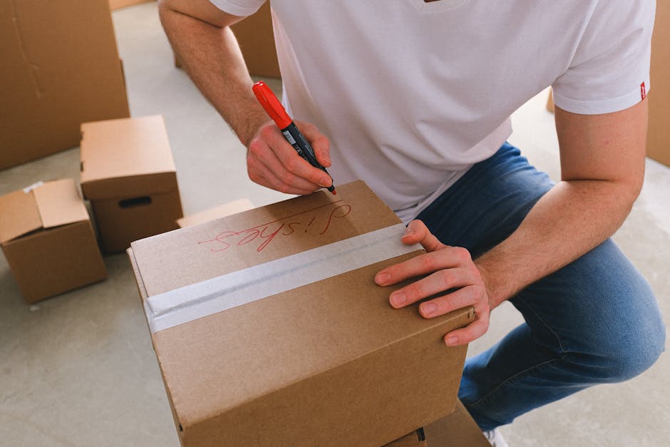 A person wearing a white t-shirt and blue jeans is kneeling indoors, using a red marker to write on a brown cardboard box that is sealed with white packing tape. The person is in the process of labeling the box, with the word 'sips' visible in red ink. Surrounding the individual are several other packed cardboard boxes of various sizes, some stacked and others placed on the floor, indicating a home relocation or packing process. The setting appears to be inside a house or flat, with neutral-colored walls and a door partially visible in the background. The scene emphasizes careful packing and labeling, which are essential steps in furniture transport and home removals, and reflects the meticulous preparation involved in professional moving services such as those offered by Man with Van Addiscombe, especially when arranging for permits with Croydon Council for effective house removals.