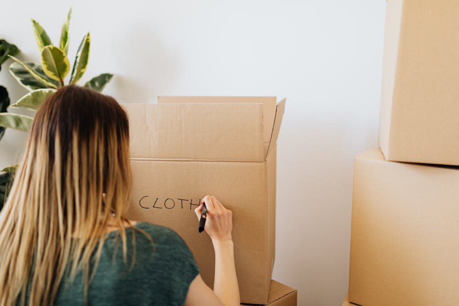 A woman with long, straight blonde hair is inside a residential property, standing in front of a large cardboard box with the word 'CLOTH' written on it in black marker. She is holding a marker in her