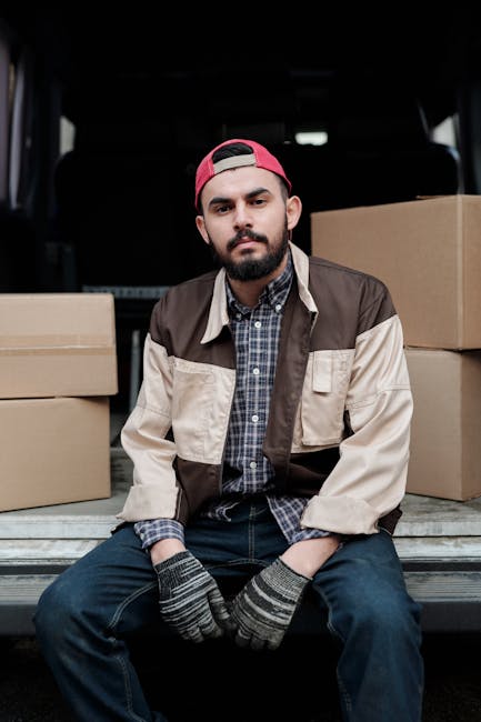 A young man with a beard, wearing a beige and brown jacket, a blue checkered shirt, a red backward cap, and grey gloves, sitting on the edge of an open van's loading area. Behind him are several large