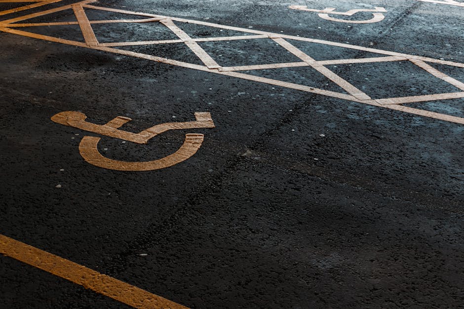 A close-up view of an asphalt parking area with white painted lines designating parking spaces, including a clearly visible yellow wheelchair accessible symbol painted on the ground. The asphalt surface appears slightly textured and weathered. In the background, partially visible are other marked parking spaces and the edge of the pavement. The scene is likely outdoors, with natural lighting highlighting the painted markings and surface details. This setting relates to moving and relocation services by illustrating typical parking provisions used by [COMPANY_NAME], Man with Van Addiscombe, during furniture transport or home relocation activities involving vehicle loading and unloading at property entrances or designated parking zones.