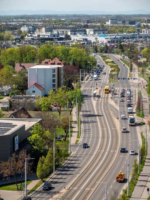 An aerial view of Lower Addiscombe Road showing a wide, multi-lane street with moving vehicles including cars, buses, and a van, flanked by green trees and residential buildings on one side and commercial properties in the distance. The road features painted lane markings, bus stops, and street lighting, with a clear sky overhead. This scene illustrates urban traffic flow typical of the area, as part of a cityscape where house removals and moving logistics might take place, supported by companies like Man with Van Addiscombe known for efficient furniture transport and home relocation services.