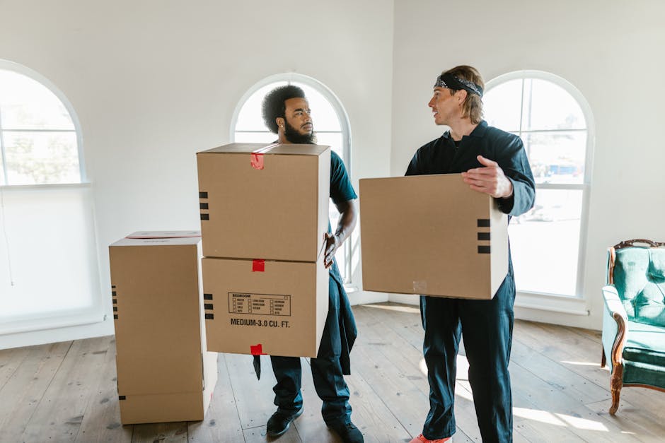 A man with dark curly hair and a beard, dressed in a navy blue t-shirt with a small logo on the chest and dark trousers, is inside a bright, empty room with light-colored walls and wooden flooring. He is holding a medium-sized cardboard box with red tape around it in his right hand while standing next to two more stacked cardboard boxes, one of which has black tape. Behind him, a large arched window allows natural light to illuminate the space, creating shadows on the floor. The scene depicts the process of packing or unpacking during a home relocation, with furniture or other moving materials not visible. This image appears to be associated with a professional removals service such as Man with Van Addiscombe, providing local moving and furniture transport solutions, as suggested by the context of house removals and the environment shown.