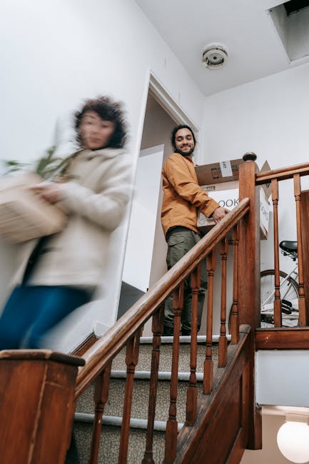Indoor scene showing two movers from Man with Van Addiscombe engaged in the home relocation process. One mover, a man with a beard wearing a brown jacket, is standing at the bottom of a wooden staircase, holding packs of boxes, likely preparing to load or unload items. The other mover, a woman dressed in a light-colored hoodie, is walking past the staircase carrying a cardboard box, with motion blur indicating movement. The staircase has wooden railings and beige carpeted steps, leading up to a doorway or landing. The corridor wall is painted white, with a smoke detector affixed to the ceiling. Multiple cardboard boxes, some labeled, are visible on the landing and near the staircase, along with packing materials. The environment suggests an interior of a residential property during a packing and moving operation, with natural lighting illuminating the scene. Man with Van Addiscombe specializes in furniture transport and packing services, assisting clients in safely managing stairway navigation during relocations.