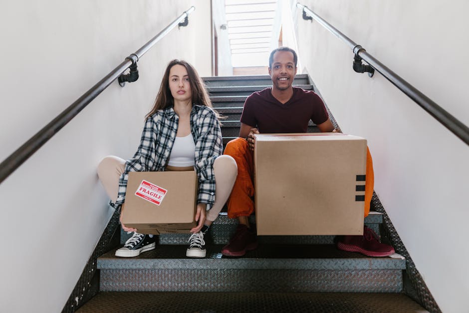 Indoor scene showing two movers from Man with Van Addiscombe engaged in the home relocation process. One mover, a man with a beard wearing a brown jacket, is standing at the bottom of a wooden staircase, holding packs of boxes, likely preparing to load or unload items. The other mover, a woman dressed in a light-colored hoodie, is walking past the staircase carrying a cardboard box, with motion blur indicating movement. The staircase has wooden railings and beige carpeted steps, leading up to a doorway or landing. The corridor wall is painted white, with a smoke detector affixed to the ceiling. Multiple cardboard boxes, some labeled, are visible on the landing and near the staircase, along with packing materials. The environment suggests an interior of a residential property during a packing and moving operation, with natural lighting illuminating the scene. Man with Van Addiscombe specializes in furniture transport and packing services, assisting clients in safely managing stairway navigation during relocations.