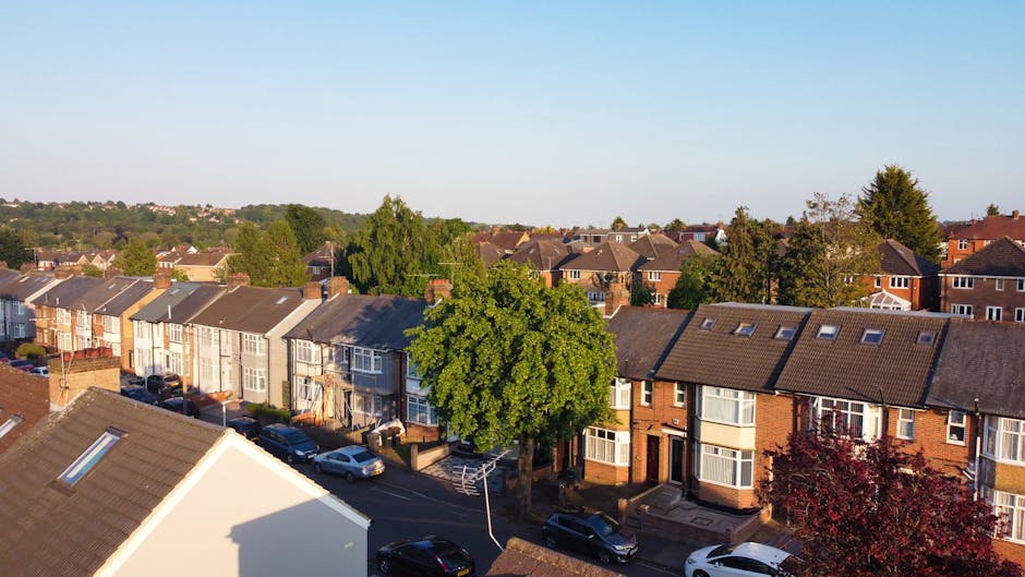 An aerial view of a residential street in Addiscombe during daylight, showing a row of terraced houses with various brick and painted facades, some with bay windows and small front gardens. Several cars are parked along the narrow street, which runs parallel to the pavement. Prominent in the foreground is a white house with a gabled roof, partially casting a shadow on the road. In the middle ground, a large, leafy green tree stands beside the sidewalk, providing natural contrast to the built environment. The background extends to additional residential buildings and greenery, under a clear blue sky. The image captures the typical suburban setting suitable for home relocations and furniture transport, relevant to professional removals services like those offered by Man with Van Addiscombe, focusing on packing, loading, and transportation processes involved in house removals.