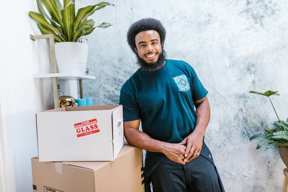 A smiling man with curly black hair and a beard, wearing a blue t-shirt with a pocket, stands indoors next to a stack of cardboard boxes used for packing. One box has a red label reading 'GLASS' and is partially open, revealing packing materials inside. The man is positioned near a white wall with a small shelf holding a potted plant, a metallic decorative object, and a blue container. Behind him, there are more indoor plants, and the environment suggests a home setting. The scene depicts packing and moving preparation, possibly part of a home relocation or furniture transport process, with clear visibility of packaging materials, boxes, and the interior space suitable for a professional removals service such as Man with Van Addiscombe.