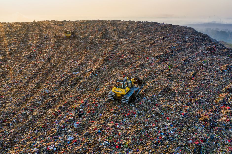 An aerial view of a large area covered in mixed waste debris, including plastic, paper, and fabric materials. In the center, a yellow and gray excavator with a long arm and bucket attachment is positi